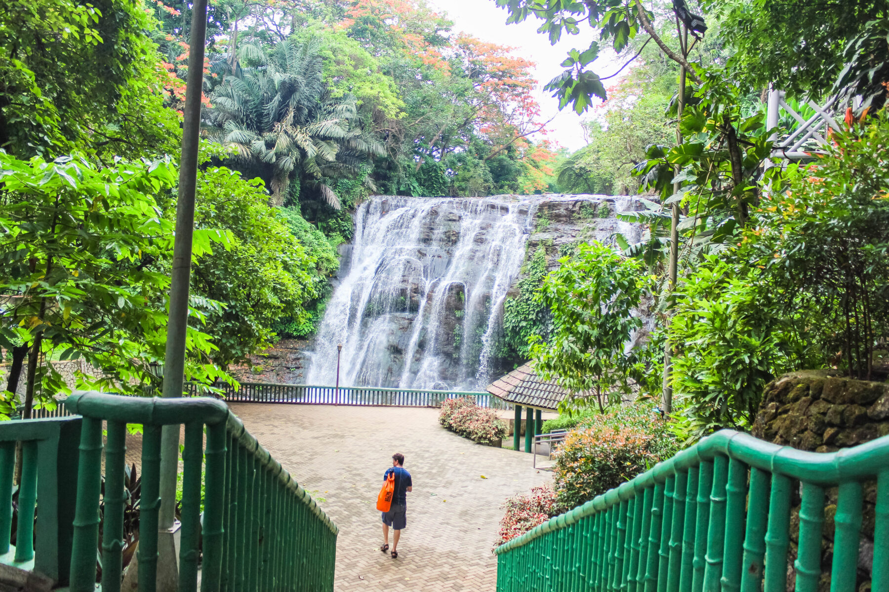 View of Hinulugang Taktak waterfall in Antipolo, with the paved viewing area and green railing in the foreground, surrounded by lush trees and tropical foliage.