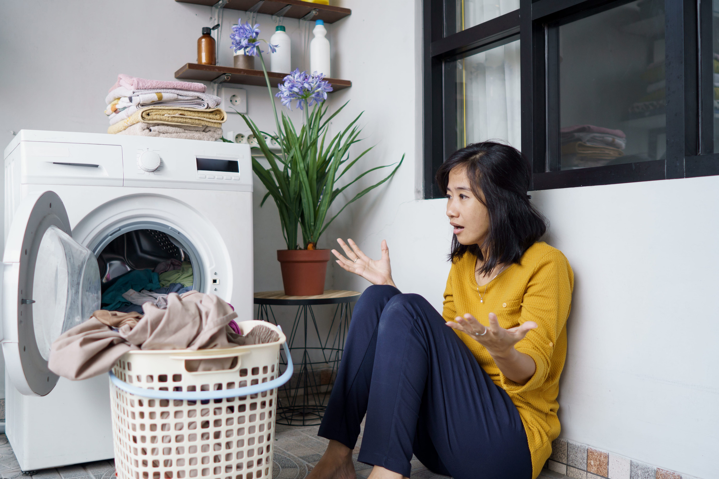 Woman gesturing beside an open washing machine and full laundry basket, illustrating frustration in a ningas cogon mentality article.